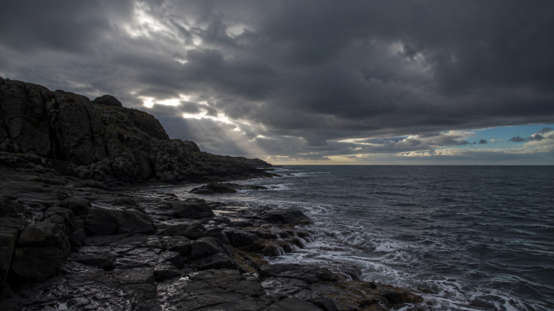 Rugged rocky shoreline under dark storm clouds with a narrow sunbeam shining over the ocean.